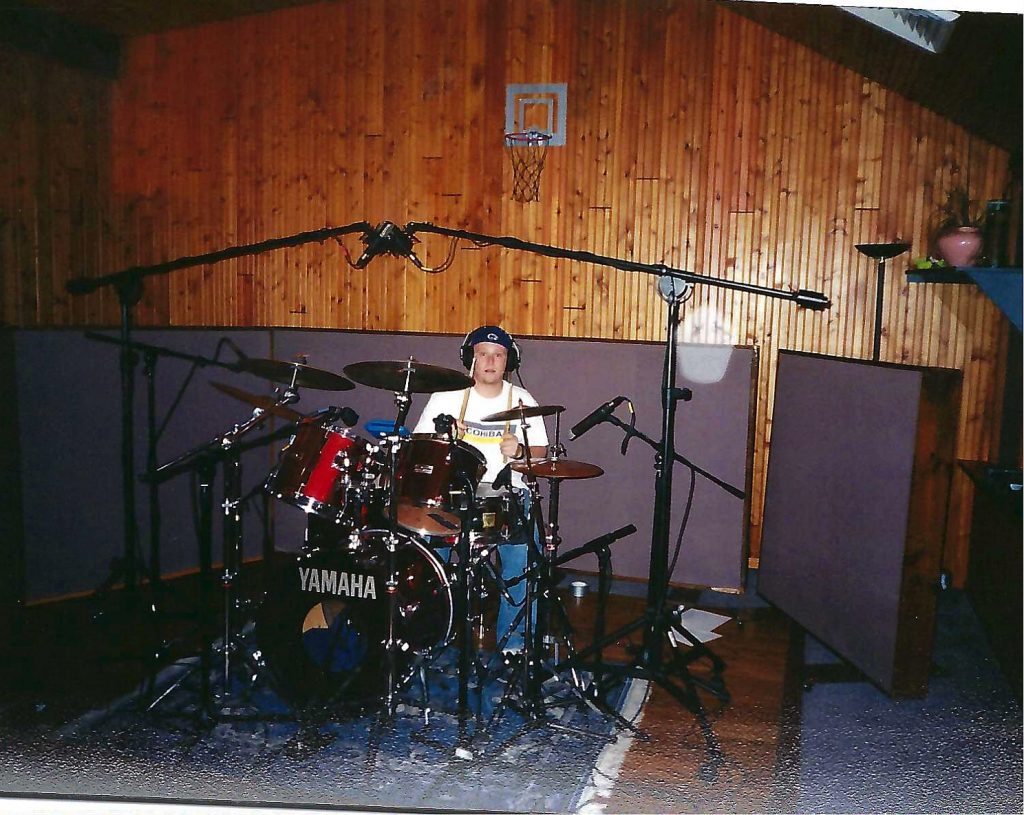 Drummer playing a red Yamaha kit in a wood-paneled recording studio, surrounded by microphones and sound baffles, early 2000s